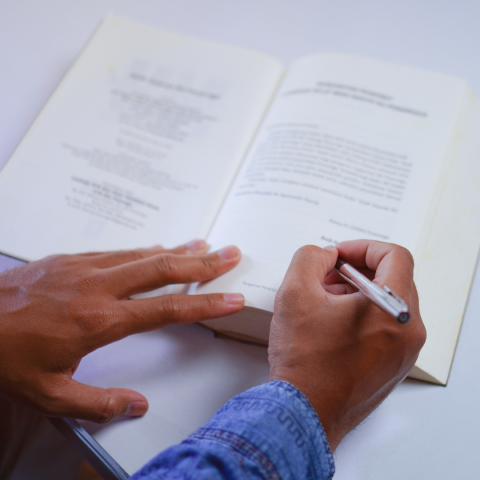 A person's hands holding an open book and writing with a pen on a page.